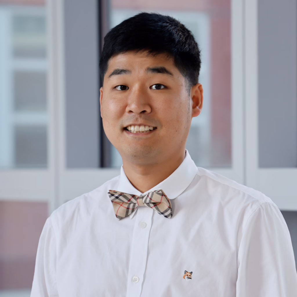Portrait of a young man in a white dress shirt with a beige plaid bow tie, smiling indoors; small fox emblem on the shirt.