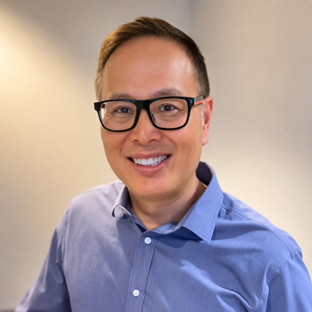 Smiling man with short hair and black glasses in a light blue button-down shirt, looking at the camera.