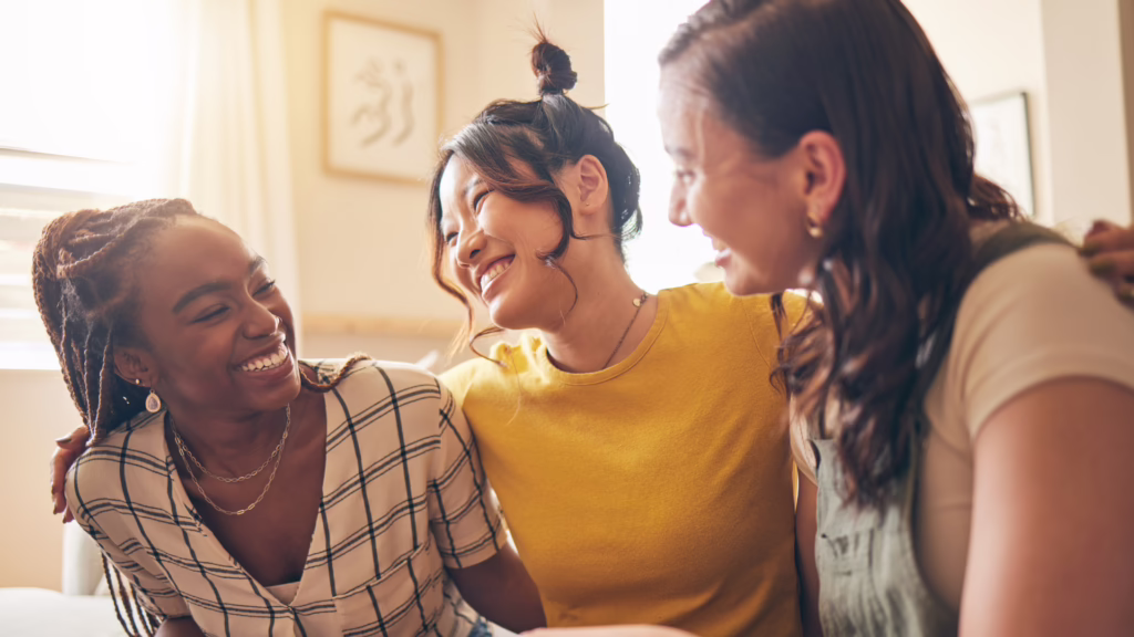 Three women smiling and leaning in toward each other in a sunlit room, sharing a joyful moment together on a casual day.